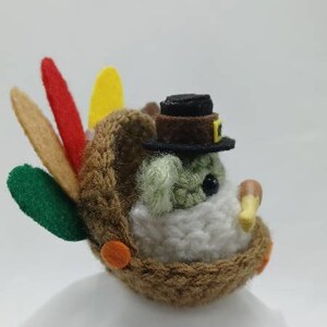 Puede incluir: Un peque&ntilde;o juguete de peluche de pavo con un sombrero de fieltro marr&oacute;n y un cuerpo de fieltro blanco. El pavo tiene plumas de cola de fieltro verde, amarillo y rojo y detalles de fieltro naranja.