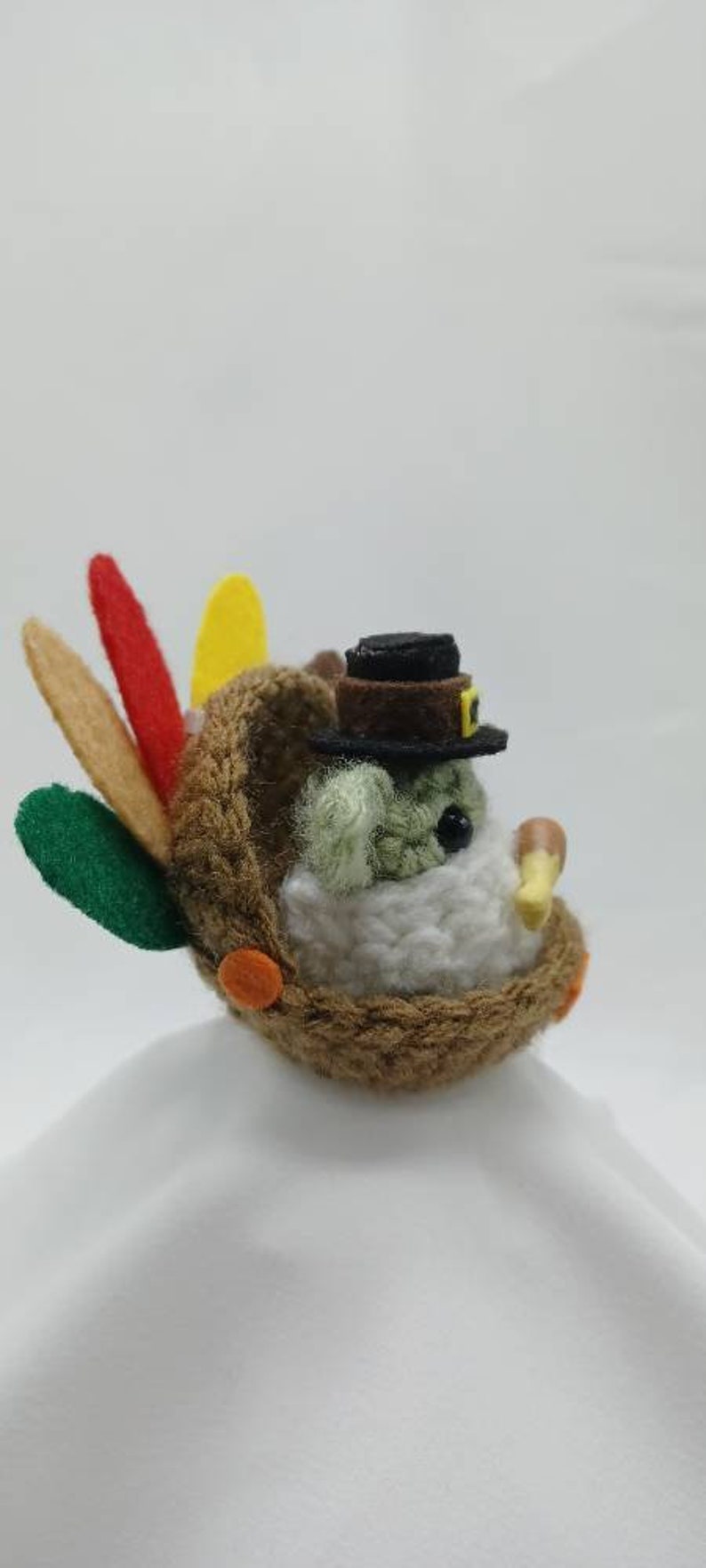 Puede incluir: Un peque&ntilde;o juguete de peluche de pavo con un sombrero de fieltro marr&oacute;n y un cuerpo de fieltro blanco. El pavo tiene plumas de cola de fieltro verde, amarillo y rojo y detalles de fieltro naranja.