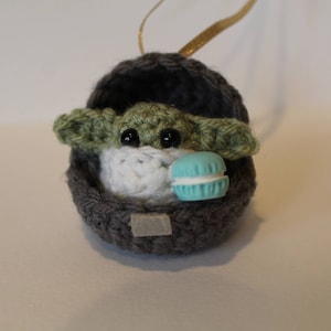 K&ouml;nnte beinhalten: Ein geh&auml;keltes Yoda-Ornament in Gr&uuml;n und Grau, das ein blaues Makron h&auml;lt. Das Ornament h&auml;ngt an einem goldenen Band.