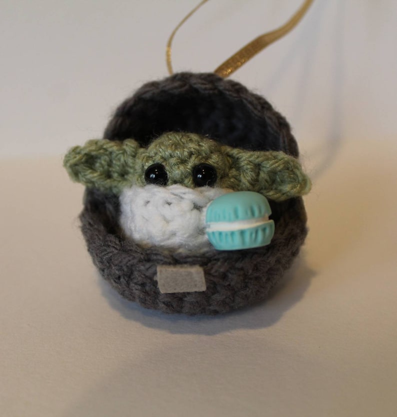 K&ouml;nnte beinhalten: Ein geh&auml;keltes Yoda-Ornament in Gr&uuml;n und Grau, das ein blaues Makron h&auml;lt. Das Ornament h&auml;ngt an einem goldenen Band.
