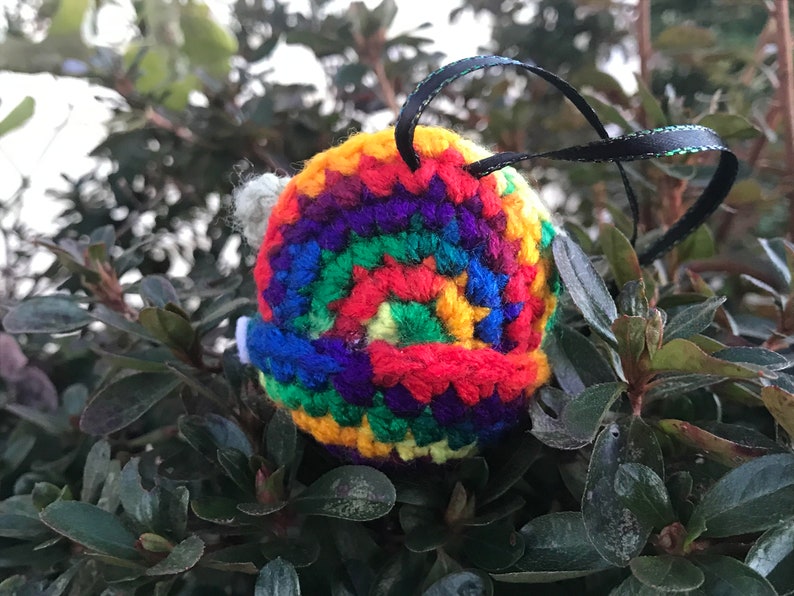 May include: A rainbow crocheted ornament with a black ribbon attached. The ornament is a spiral of colorful yarn.