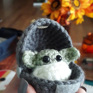 May include: A crocheted green baby Yoda doll in a gray crocheted cradle. The doll has black eyes and a white crocheted blanket.