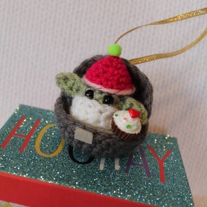 May include: A crocheted Yoda ornament wearing a red and white hat and holding a cupcake. The ornament is hanging from a gold ribbon and is sitting on a gift box with a blue glitter top and a pink and white polka dot bottom.