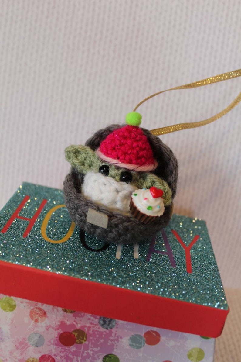 May include: A crocheted Yoda ornament wearing a red and white hat and holding a cupcake. The ornament is hanging from a gold ribbon and is sitting on a gift box with a blue glitter top and a pink and white polka dot bottom.