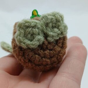Peut inclure: Un petit objet rond en crochet marron avec deux accents circulaires textur&eacute;s vert clair sur la surface sup&eacute;rieure. Un minuscule d&eacute;tail vert vif et jaune est visible au centre, et une petite extension en forme de feuille verte d&eacute;passe sur le c&ocirc;t&eacute;.