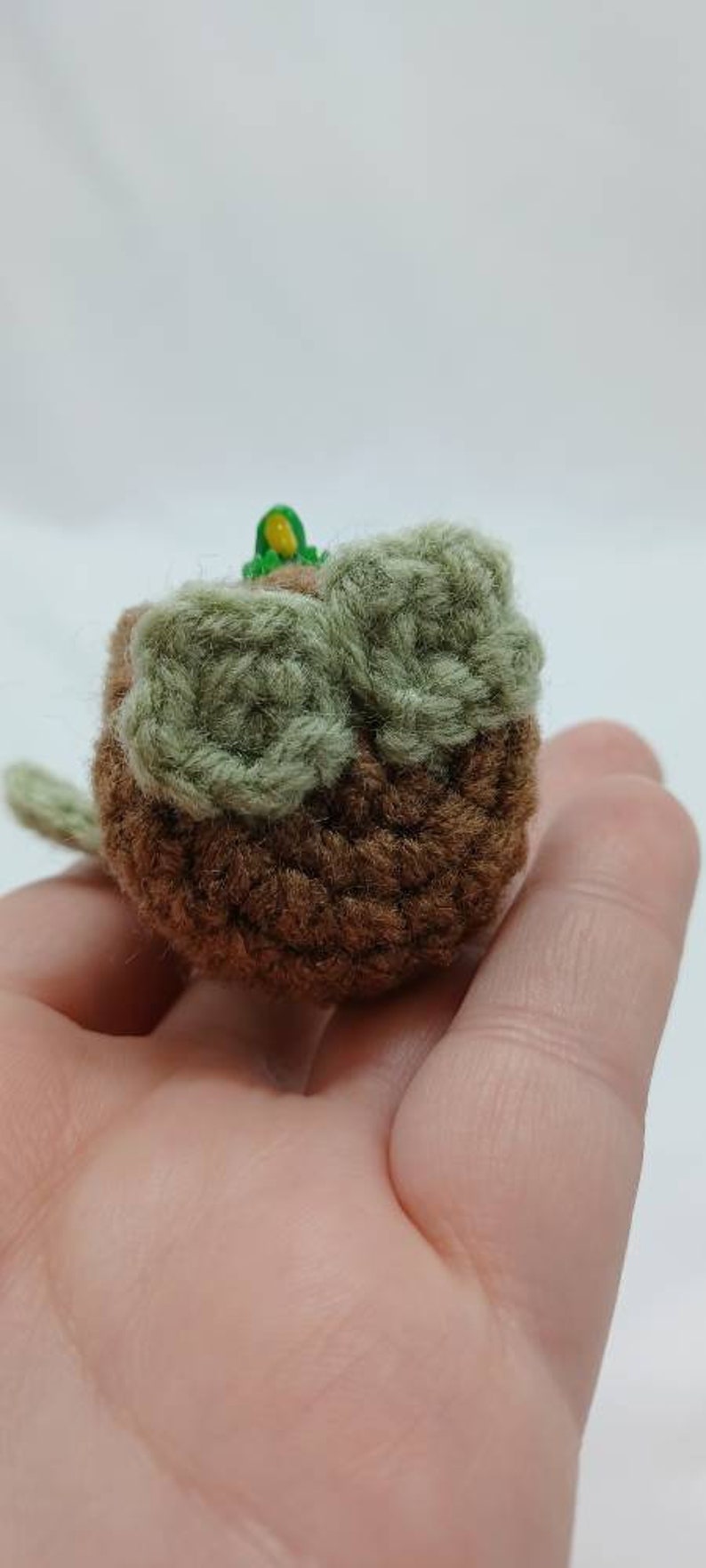 Peut inclure: Un petit objet rond en crochet marron avec deux accents circulaires textur&eacute;s vert clair sur la surface sup&eacute;rieure. Un minuscule d&eacute;tail vert vif et jaune est visible au centre, et une petite extension en forme de feuille verte d&eacute;passe sur le c&ocirc;t&eacute;.