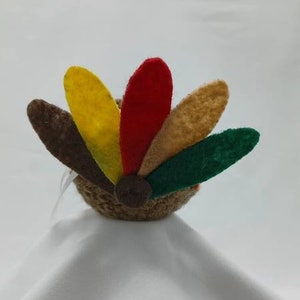 Puede incluir: Un pavo de fieltro marr&oacute;n con una cola de fieltro de colores. La cola tiene plumas rojas, amarillas, verdes y marrones.