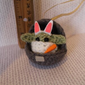 May include: A crocheted Yoda doll wearing a pink bunny ear headband and holding a carrot. The doll is sitting in a gray crocheted basket.