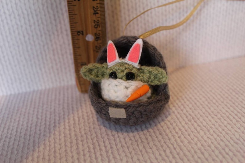 May include: A crocheted Yoda doll wearing a pink bunny ear headband and holding a carrot. The doll is sitting in a gray crocheted basket.