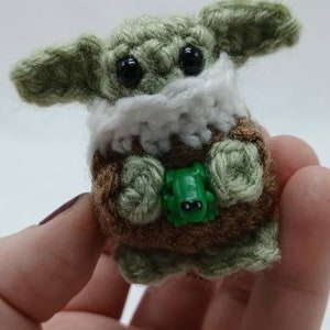 Peut inclure: Une petite cr&eacute;ature crochet&eacute;e &agrave; la peau vert clair, aux grands yeux noirs et aux oreilles pro&eacute;minentes. Elle porte un v&ecirc;tement marron avec un col blanc et tient une minuscule grenouille en plastique vert vif entre ses mains en laine.