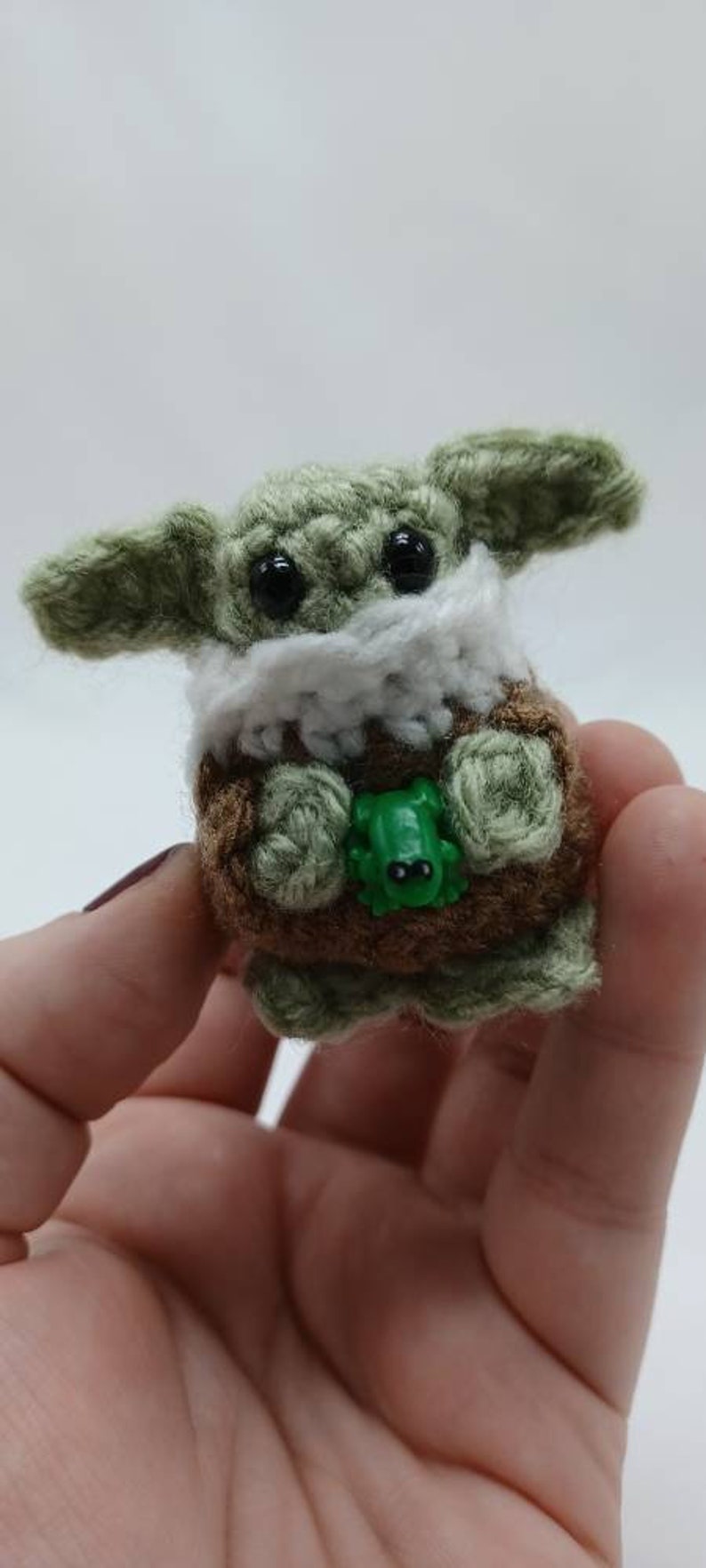 Peut inclure: Une petite cr&eacute;ature crochet&eacute;e &agrave; la peau vert clair, aux grands yeux noirs et aux oreilles pro&eacute;minentes. Elle porte un v&ecirc;tement marron avec un col blanc et tient une minuscule grenouille en plastique vert vif entre ses mains en laine.