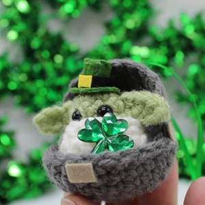 May include: A small crocheted Yoda doll wearing a green hat with a yellow buckle, holding a green shamrock. The doll is sitting in a grey crocheted egg-shaped container.