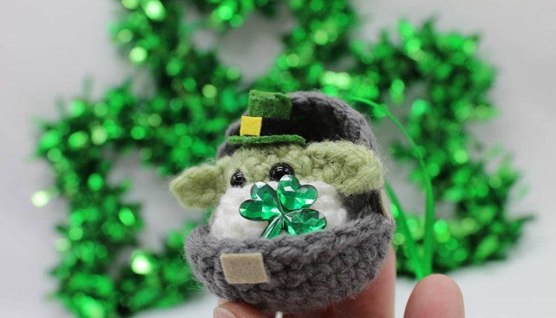 May include: A small crocheted Yoda doll wearing a green hat with a yellow buckle, holding a green shamrock. The doll is sitting in a grey crocheted egg-shaped container.