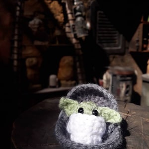 May include: A small crocheted plush toy of a green alien character, known as Baby Yoda, sitting in a gray crocheted pod. The toy is sitting on a wooden surface.