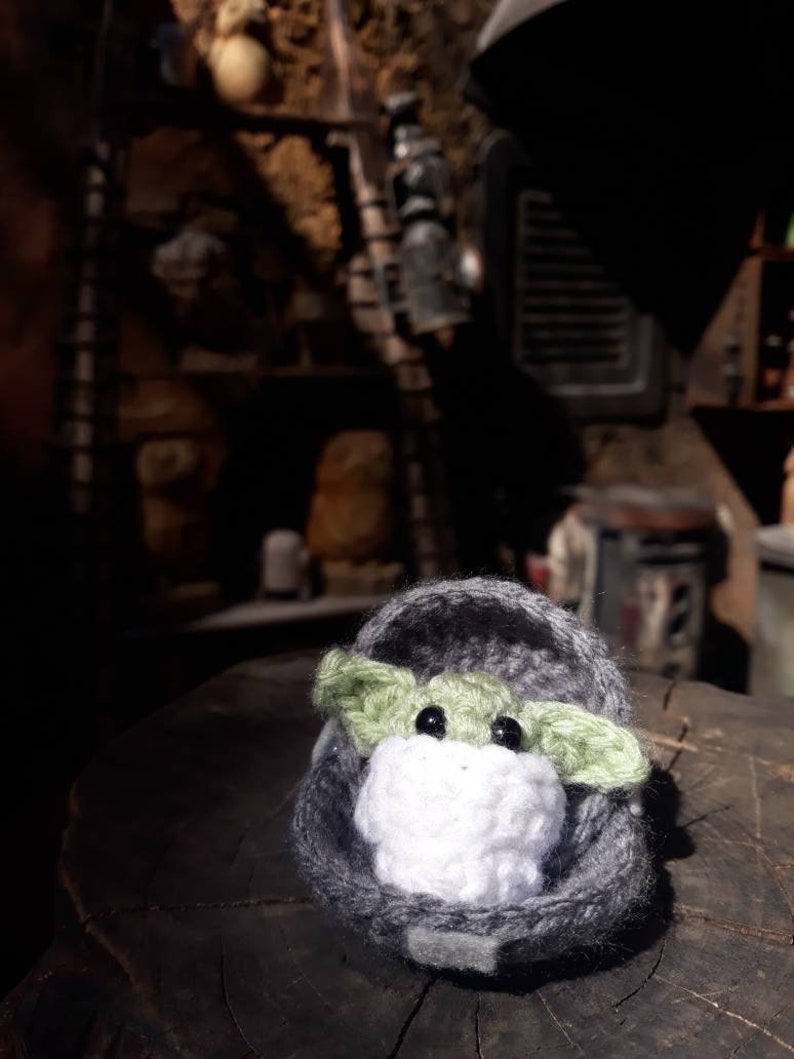 May include: A small crocheted plush toy of a green alien character, known as Baby Yoda, sitting in a gray crocheted pod. The toy is sitting on a wooden surface.