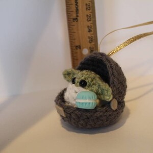 K&ouml;nnte beinhalten: Ein geh&auml;keltes Ornament eines gr&uuml;nen Baby-Alien-Charakters, der ein blaues und wei&szlig;es Makron h&auml;lt. Der Charakter befindet sich in einer grauen geh&auml;kelten Wiege mit einem goldenen Band.