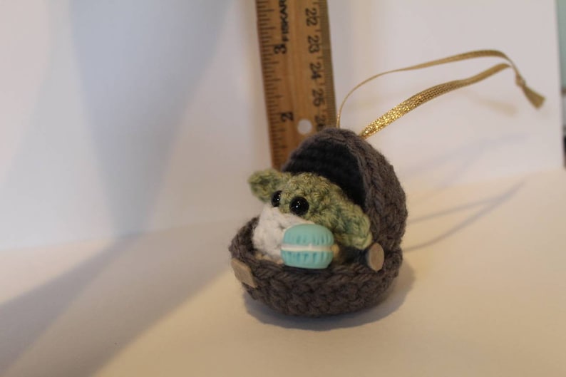 K&ouml;nnte beinhalten: Ein geh&auml;keltes Ornament eines gr&uuml;nen Baby-Alien-Charakters, der ein blaues und wei&szlig;es Makron h&auml;lt. Der Charakter befindet sich in einer grauen geh&auml;kelten Wiege mit einem goldenen Band.