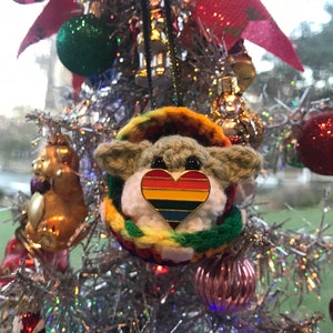 May include: A crocheted Yoda ornament with a rainbow heart pin hanging on a silver Christmas tree. The ornament is surrounded by other Christmas ornaments, including a gold bear and a red glitter ball.
