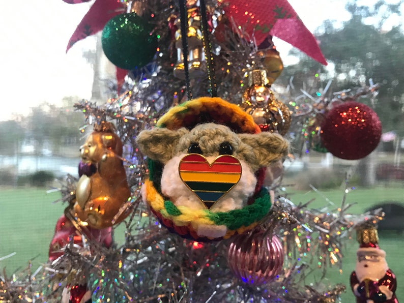 May include: A crocheted Yoda ornament with a rainbow heart pin hanging on a silver Christmas tree. The ornament is surrounded by other Christmas ornaments, including a gold bear and a red glitter ball.