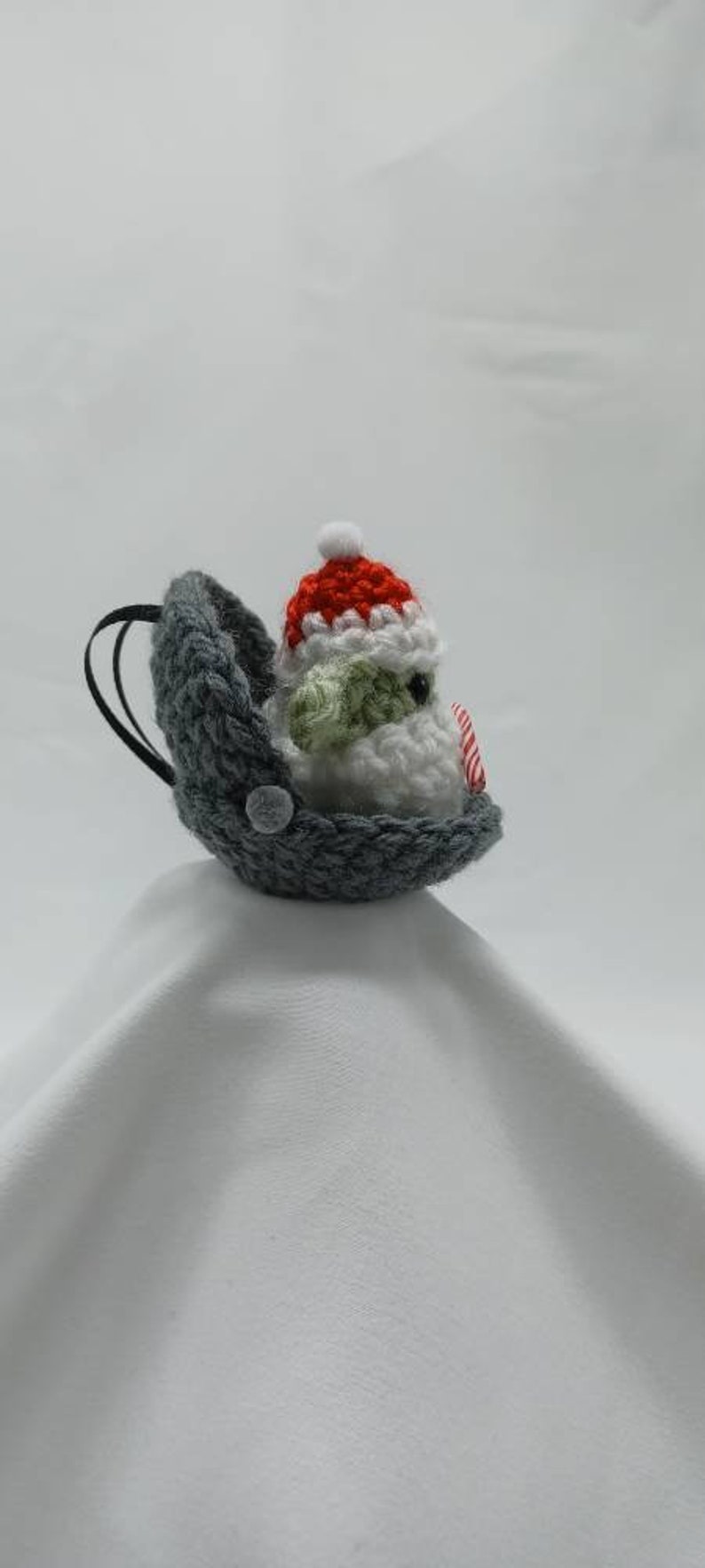 May include: A gray crocheted stocking with a white crocheted snowman wearing a red and white crocheted hat. The snowman is holding a red and white candy cane.
