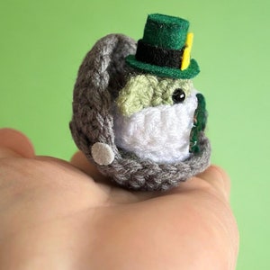 May include: A small crocheted Yoda wearing a green leprechaun hat with a yellow buckle. The Yoda is sitting in a grey crocheted pod.