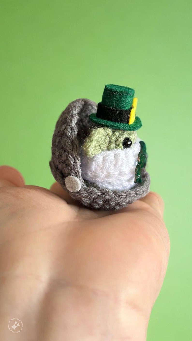 May include: A small crocheted Yoda wearing a green leprechaun hat with a yellow buckle. The Yoda is sitting in a grey crocheted pod.