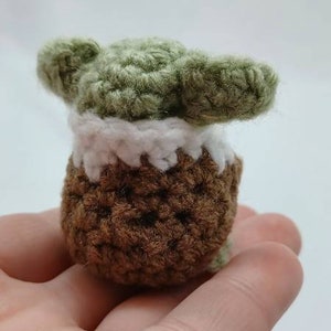Peut inclure: Une petite figurine crochet&eacute;e &agrave; la main pr&eacute;sentant une t&ecirc;te verte avec de grandes oreilles pointues, un col blanc et un corps marron textur&eacute;. L'objet est fabriqu&eacute; en fil avec des mailles visibles, affichant une forme compacte et arrondie.