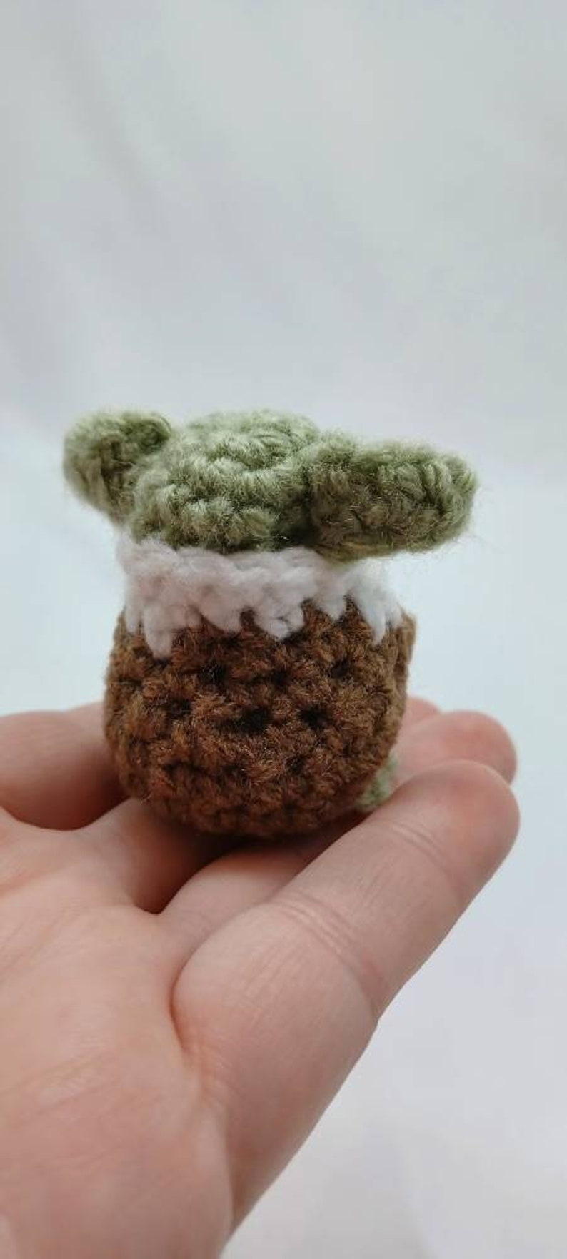 Peut inclure: Une petite figurine crochet&eacute;e &agrave; la main pr&eacute;sentant une t&ecirc;te verte avec de grandes oreilles pointues, un col blanc et un corps marron textur&eacute;. L'objet est fabriqu&eacute; en fil avec des mailles visibles, affichant une forme compacte et arrondie.