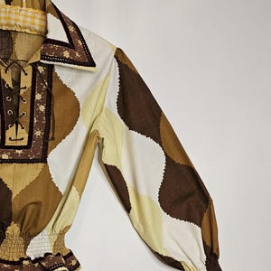 May include: A brown, white, and yellow patterned long-sleeve shirt with a lace-up neckline and elastic cuffs. The shirt has a brown trim with floral embroidery.