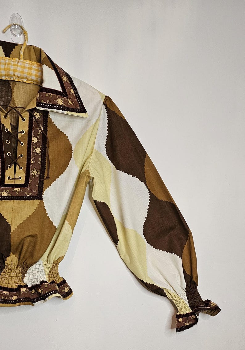 May include: A brown, white, and yellow patterned long-sleeve shirt with a lace-up neckline and elastic cuffs. The shirt has a brown trim with floral embroidery.