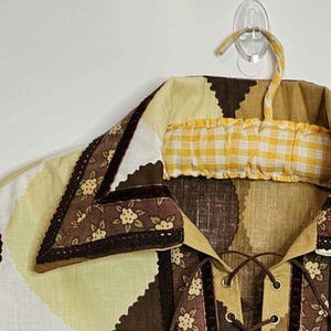 May include: A brown and yellow patterned shirt with a lace-up front and a brown velvet trim. The shirt has a collar with a brown and white floral pattern and a yellow and white checkered fabric insert.
