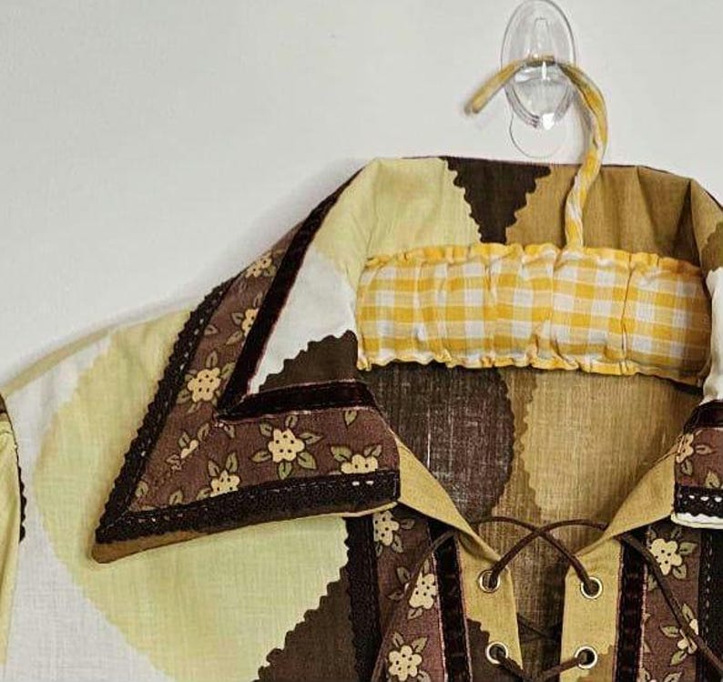 May include: A brown and yellow patterned shirt with a lace-up front and a brown velvet trim. The shirt has a collar with a brown and white floral pattern and a yellow and white checkered fabric insert.