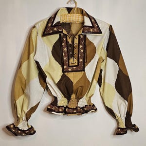 May include: A brown, white, and yellow patterned blouse with a lace-up neckline and a ruffled trim. The blouse has long sleeves with elastic cuffs and a gathered waist.