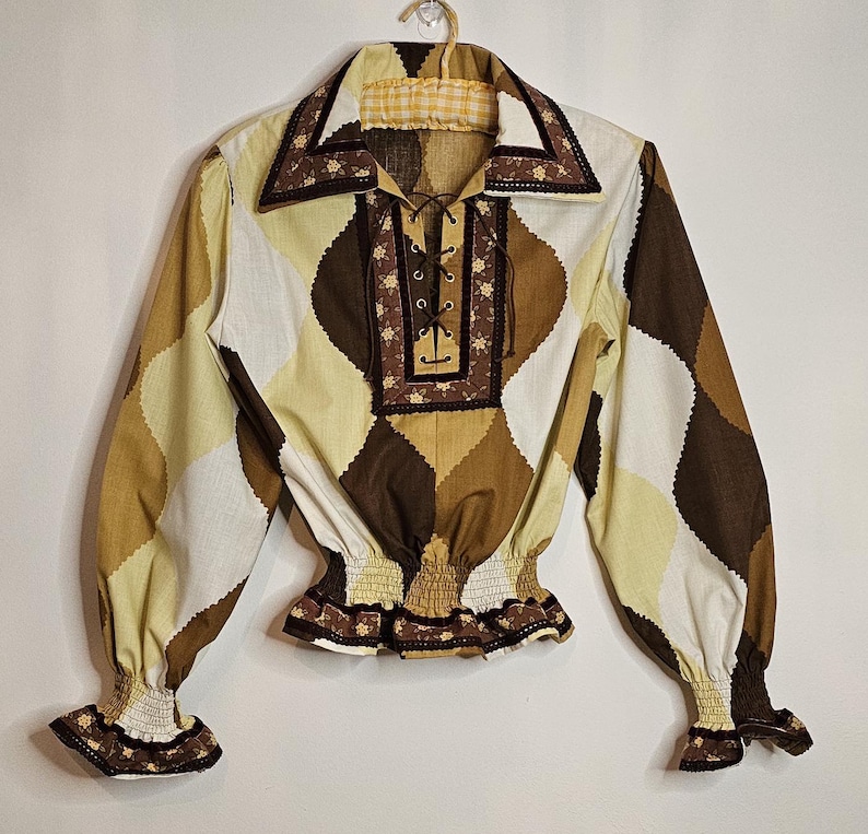 May include: A brown, white, and yellow patterned blouse with a lace-up neckline and a ruffled trim. The blouse has long sleeves with elastic cuffs and a gathered waist.