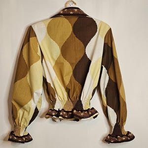 May include: A brown, white, and yellow patterned blouse with a ruffled trim and elastic waistband. The blouse has long sleeves with ruffled cuffs.