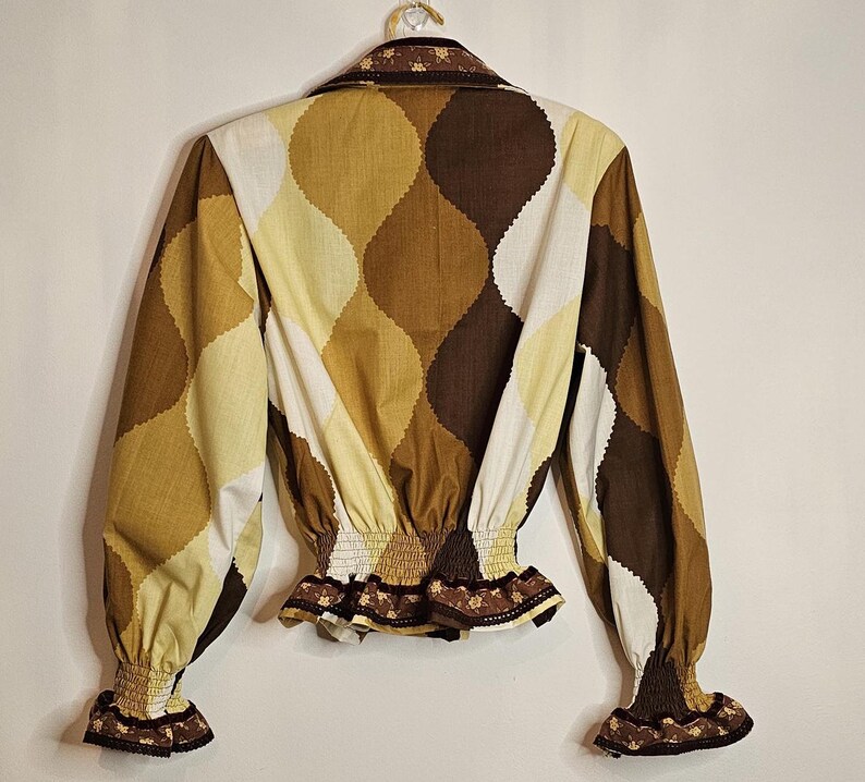 May include: A brown, white, and yellow patterned blouse with a ruffled trim and elastic waistband. The blouse has long sleeves with ruffled cuffs.