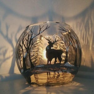 May include: A clear, round glass candle holder with a winter forest scene painted on the exterior. A silhouette of a deer stands in the center, surrounded by trees and snow. The candle inside casts a warm glow.