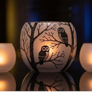 May include: A set of three glass candle holders. The central holder is a large, round vase with a black and white owl and tree design. Two smaller, round, frosted glass candle holders flank the central piece. All three are lit with a warm glow.