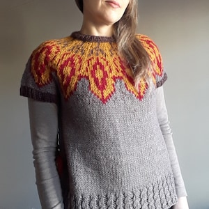 Può includere: Una donna indossa un maglione di lana marrone con un motivo geometrico colorato sul collo. Il maglione ha maniche corte e un motivo a treccia sul fondo.