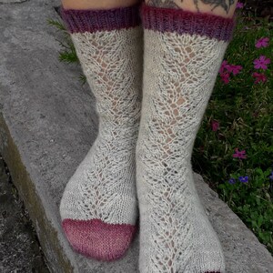 Può includere: Un paio di calze lavorate a maglia a mano con un motivo traforato di colore grigio chiaro. Le calze hanno un tallone e una punta di colore rosa e bordeaux.