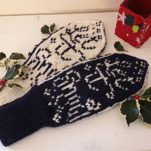 May include: Two knitted mittens, one cream and one navy blue, with a white snowflake pattern. The mittens are displayed with holly and a small red box decorated with a snowflake.