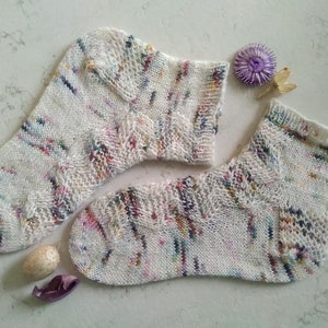 May include: A pair of white ankle socks with a colorful speckled pattern. The socks are knitted with a delicate lace design.