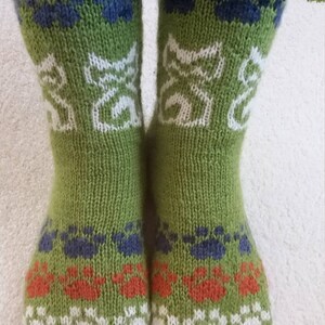 Peut inclure: Une paire de chaussettes vertes tricotées avec des empreintes de pattes et des motifs de chats. Les chaussettes présentent des empreintes de pattes blanches, orange et bleues, et des silhouettes de chats blancs. Les poignets sont côtelés.
