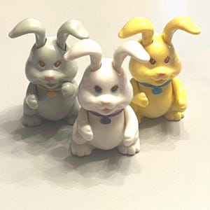 May include: Three ceramic bunny figurines in pastel colors: gray, white, and yellow. Each bunny has large ears, a round body, and a small pendant around its neck. The bunnies are sitting upright.