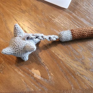 May include: A handmade crochet toy in the shape of a gray shark head connected by a chain to a brown and gray cylinder. The toy is made of soft yarn and is likely intended for a baby or toddler.
