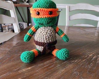 May include: A green, orange, and brown crocheted toy turtle with a black eye mask. The turtle is sitting on a wooden table.