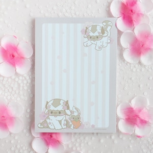 May include: A pink and white striped notepad with two cartoon illustrations of a white furry creature with brown horns and a pink flower on its head. The notepad has a light blue background with a pink and white striped pattern.