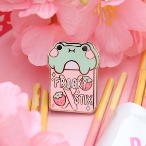 Puede incluir: Un pin de esmalte dorado con una rana verde sosteniendo una caja de "Frogy Stix" con un diseño de fresa. La caja es rosa con letras blancas y un patrón de diamantes.