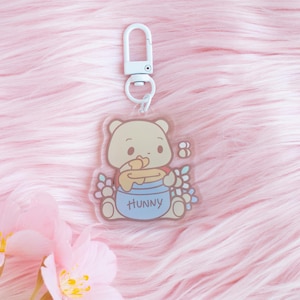May include: A clear acrylic keychain featuring a cartoon bear holding a honey pot with the word "Hunny" on it. The bear is surrounded by flowers and a small bee.