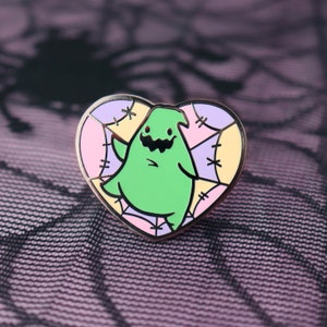 May include: A heart-shaped enamel pin featuring a green cartoon character with a jagged mouth and stitched details. The pin has a copper-colored border and is set against a patchwork background of pastel colors.